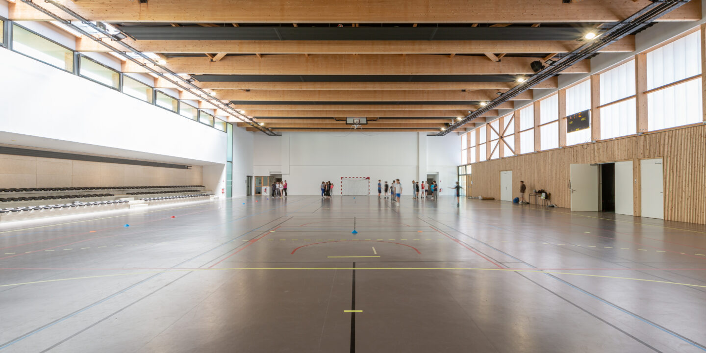Intérieur du gymnase du groupe scolaire Largenté à Bayonne avec charpente bois apparente et terrain multisports.