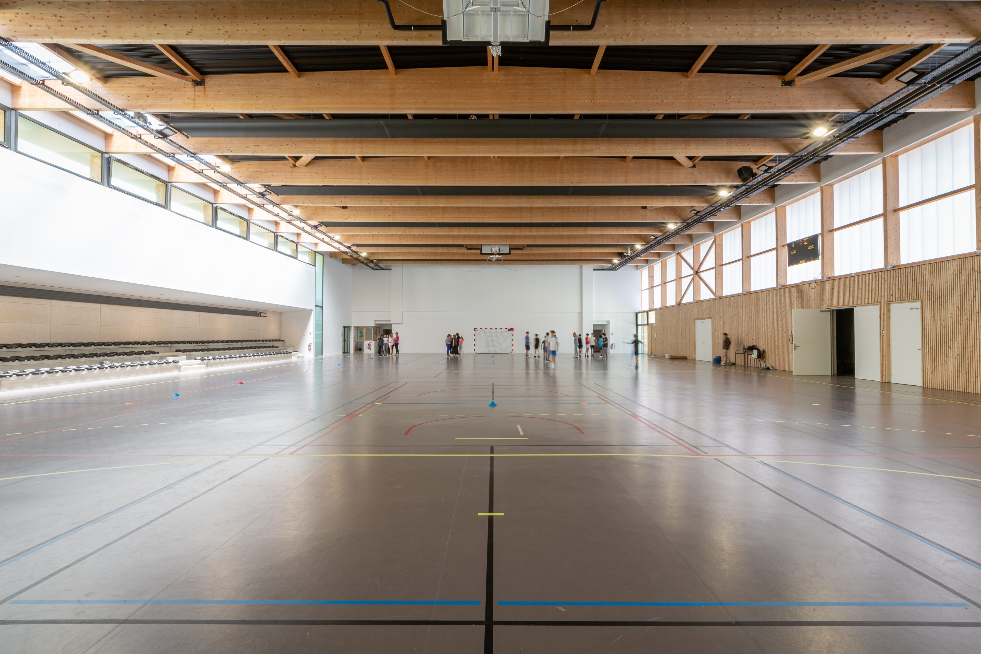 Intérieur du gymnase du groupe scolaire Largenté à Bayonne avec charpente bois apparente et terrain multisports.