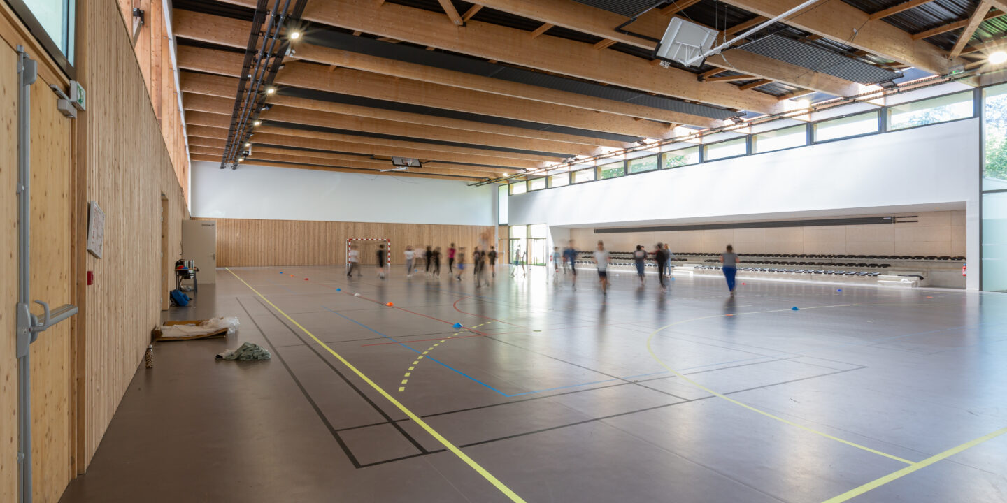 Grand volume intérieur du gymnase du groupe scolaire Largenté à Bayonne avec charpente en bois lamellé et équipements sportifs.