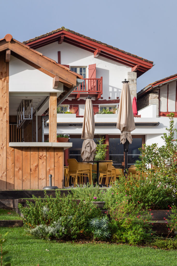 Façade arrière de l'Hôtel Oppoca à Ainhoa avec toit à pans basque, terrasse, jardin paysager et allée en pavés menant à l'entrée.