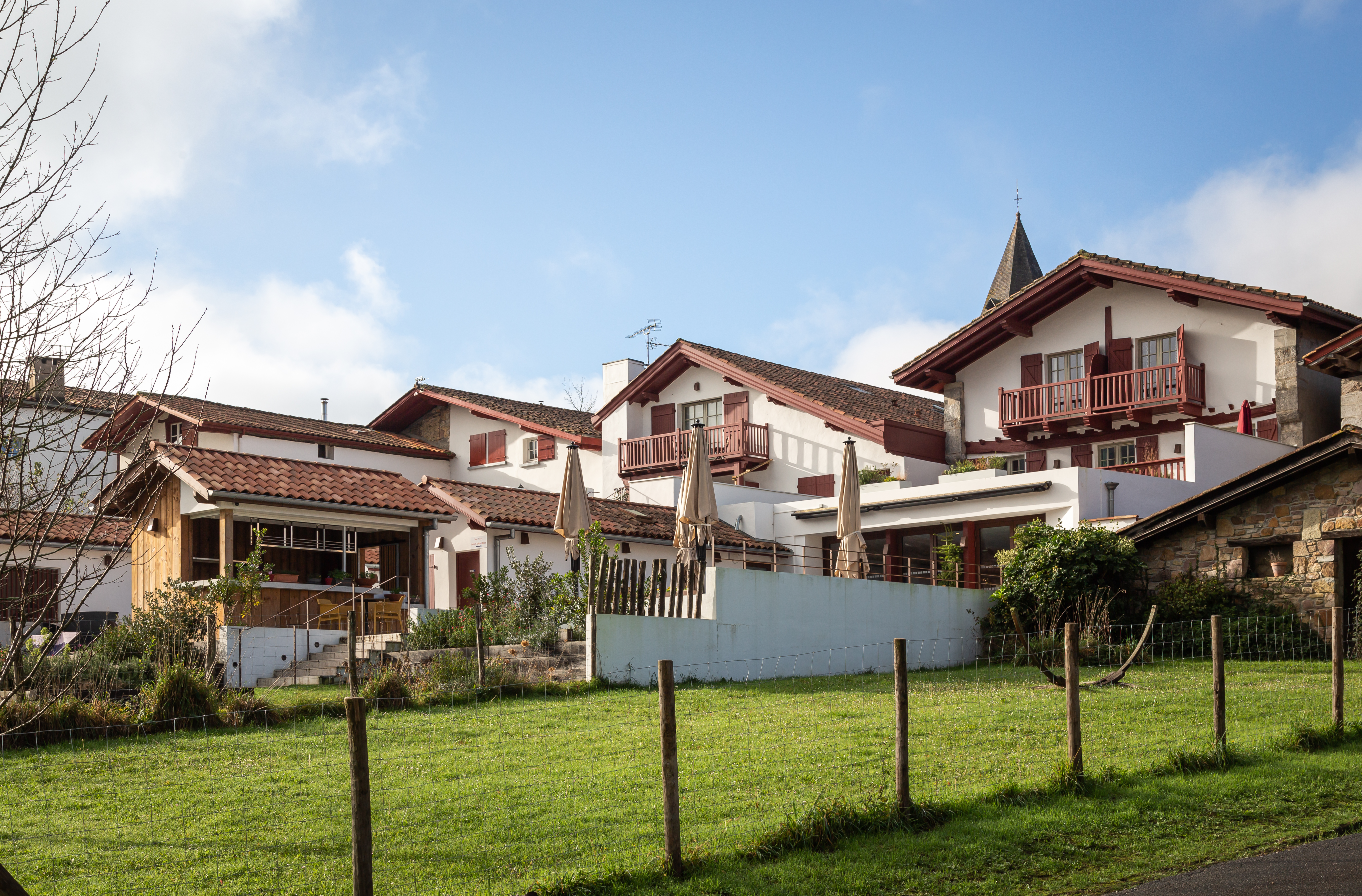Façade arrière de l'Hôtel Oppoca à Ainhoa avec toit à pans basque, terrasse, jardin paysager et allée en pavés menant à l'entrée.