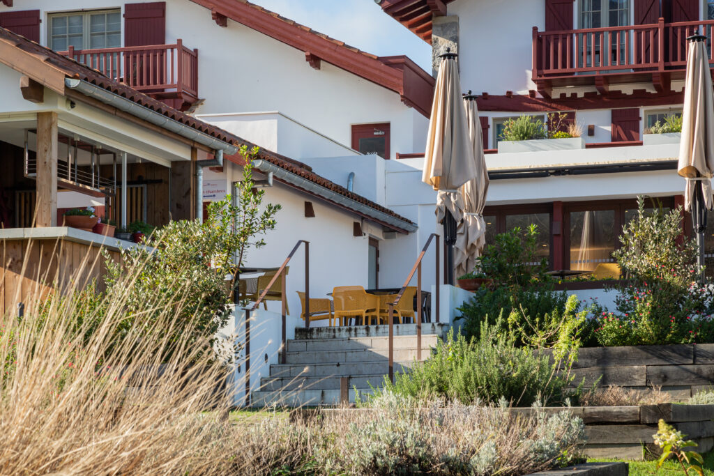 Façade arrière de l'Hôtel Oppoca à Ainhoa avec toit à pans basque, terrasse, jardin paysager et allée en pavés menant à l'entrée.