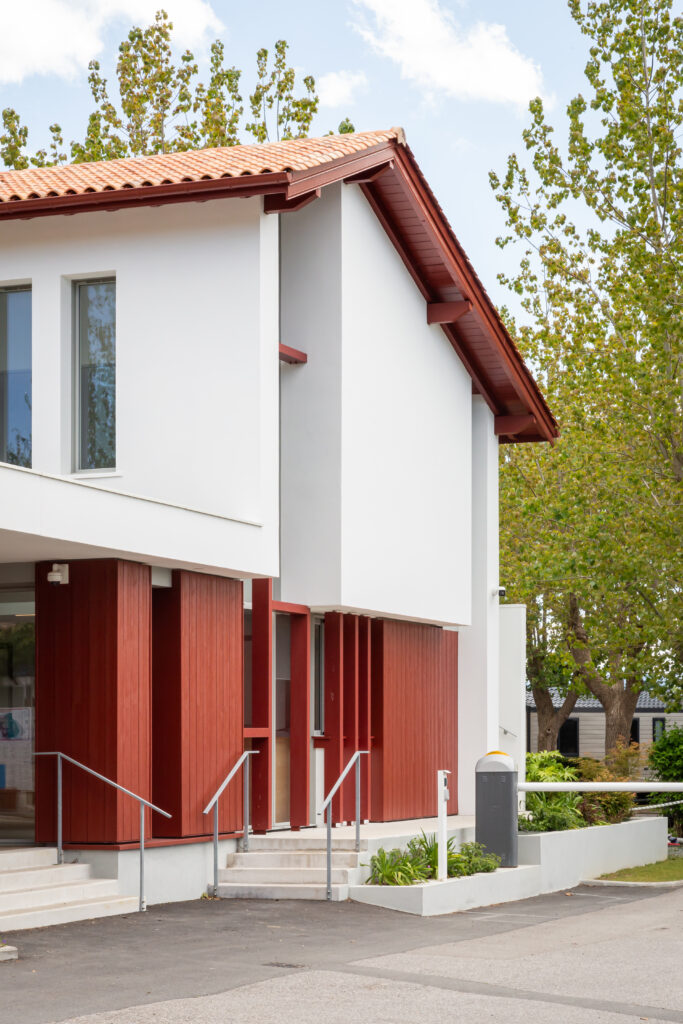 • Bâtiment d’accueil du Camping Oyam à Bidart conçu par XB Architectes, avec façade blanche, bardage bois rouge et toiture en tuiles.