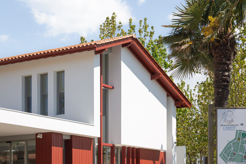 • Bâtiment d’accueil du Camping Oyam à Bidart conçu par XB Architectes, avec façade blanche, bardage bois rouge et toiture en tuiles.